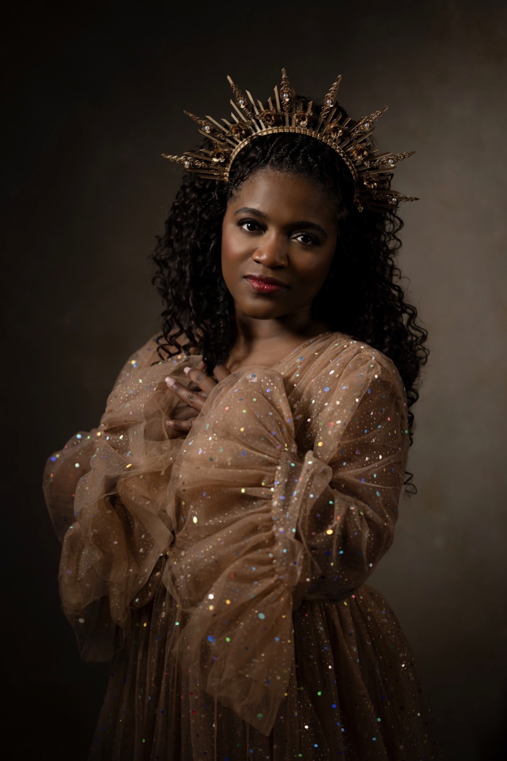 Ava Kay Rose in a sheer gold embellished gown and a sunburst crown, posed against a dark background