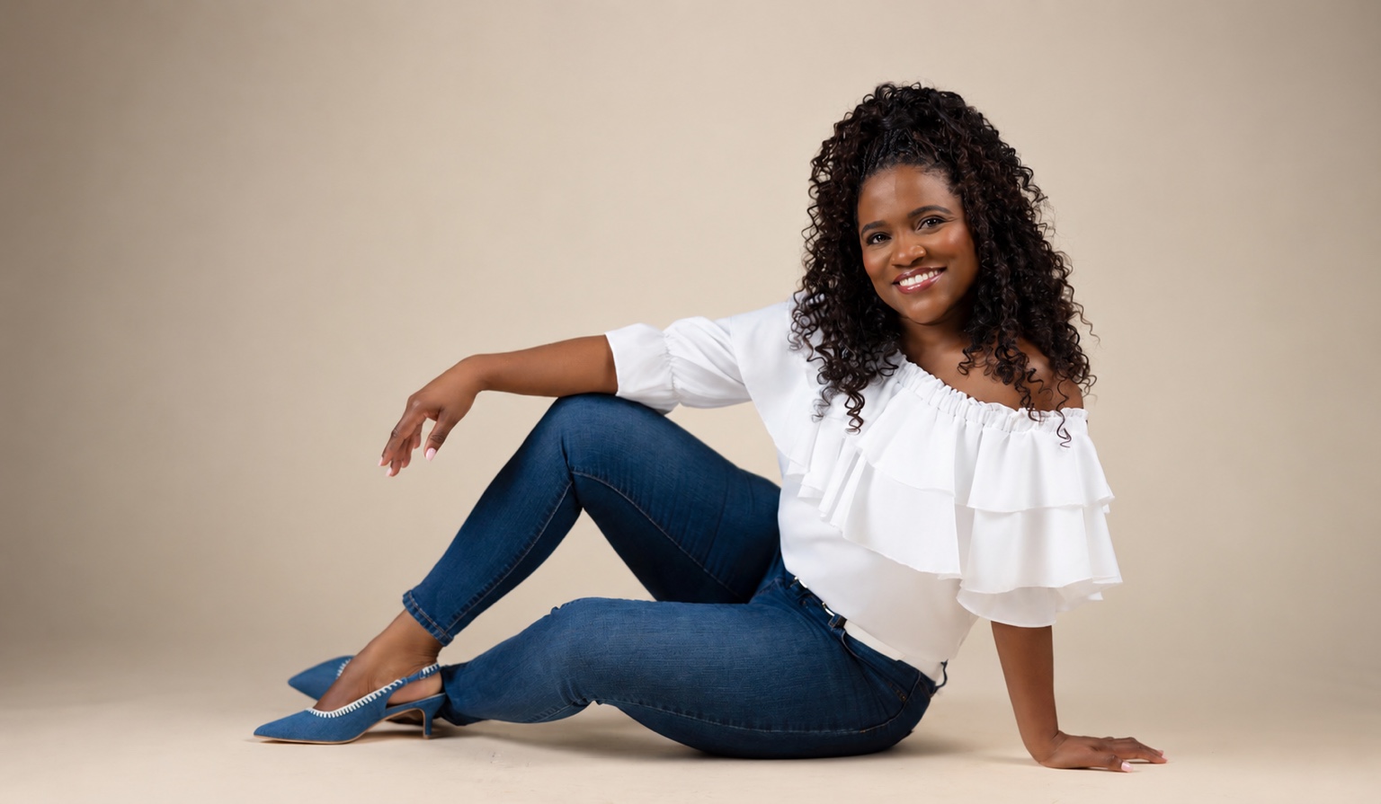 Ava Kay Rose seated on a studio floor in a white ruffled blouse, jeans, and blue heels